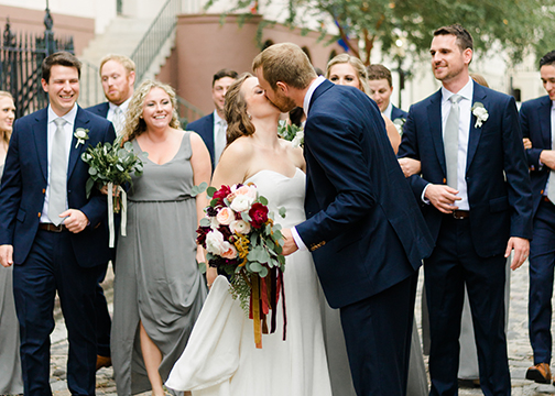 bride and groom kiss in front of bridal party holding bridal bouquets blush and red wedding flowers