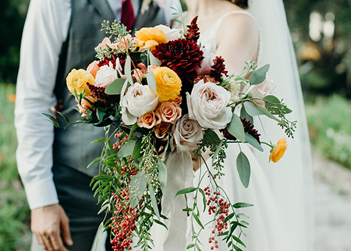 bride and groom in background holding orange garnet red and blush wedding floral